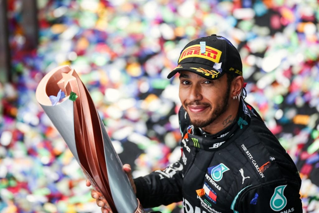 A happy Lewis Hamilton looks up and smiles at the camera holding a winners trophy