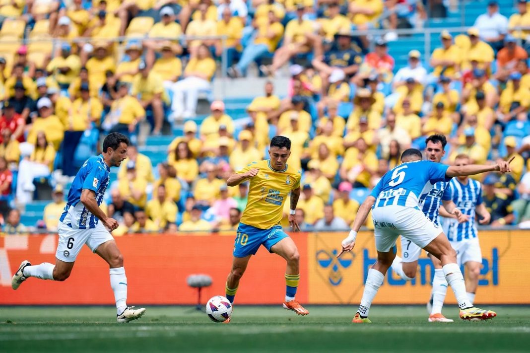 Jogador do Las Palmas conduz a bola cercado por adversários do Alavés em um confronto recente entre as equipes, válido pela LaLiga. A torcida, predominantemente vestida de amarelo, acompanha atentamente a partida no estádio.